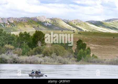 Two men fishing and a third man rowing in the Missouri River with backs to the camera. Foothills with green grasss are in the background. Stock Photo