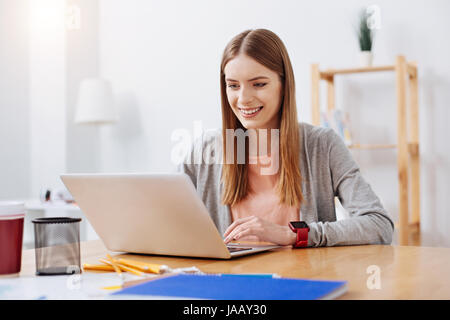 Young student. Young nice curious lady sitting at the table and typing on her computer while working on her project for upcoming exam Stock Photo