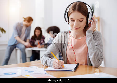 Selecting important pieces. Beautiful smart motivated lady wearing headphone and listening to audio lesson while making notes in her notebook Stock Photo