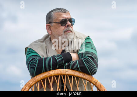 Outdoor portrait of senior man in black sunglasses leant on a wicker chair back and looking Stock Photo