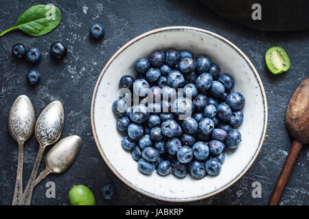 Still life of fresh blueberries in bowl, vintage cutlery, baby kiwi and spinach on stone table. Top view, closeup Stock Photo
