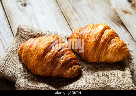 Two fresh croissants on burlap textile on rustic wooden table. Closeup view Stock Photo