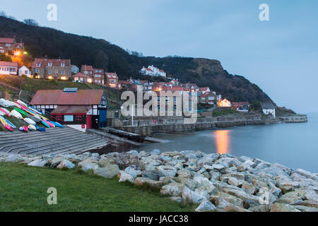 Lights are lit in houses in evening view of pretty quaint coastal village, with cliffs, boats & calm sea - Runswick Bay, North Yorkshire, England, UK. Stock Photo