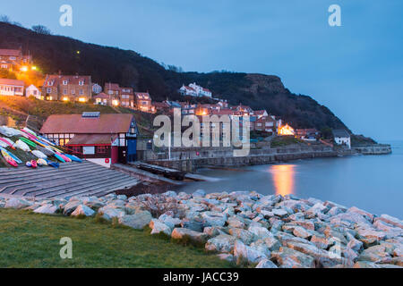 Lights are lit in houses in evening view of pretty quaint coastal village, with cliffs, boats & calm sea - Runswick Bay, North Yorkshire, England, UK. Stock Photo