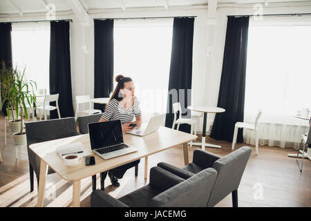 Young attractive woman is sitting at the table in a cafe with her laptop and working while looking away thoughtfully. Stock Photo