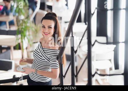 Confident team leader. Confident young woman holding a cup of hot coffee in her hands and looking at camera with smile while her colleagues working in the background. Stock Photo