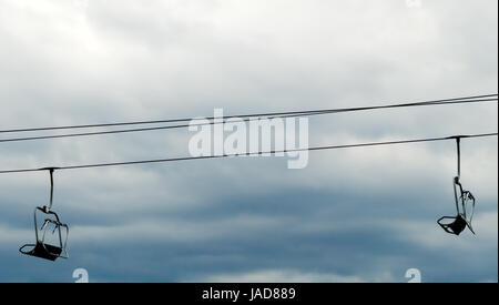 Two empty ski lift chairs hanging on steel cable against overcast sky. Stock Photo