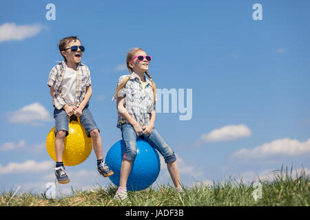 Brother and sister playing on the field at the day time. Children having fun outdoors. They jumping on inflatable balls on the lawn. Concept of friend Stock Photo