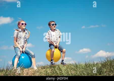 Brother and sister playing on the field at the day time. Children having fun outdoors. They jumping on inflatable balls on the lawn. Concept of friend Stock Photo