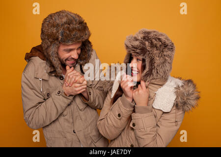 Happy couple in warm winter clothing wearing warm furry caps and jackets laughing as they stand looking at each other rubbing their hands against the cold, on an orange studio background Stock Photo