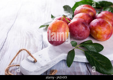 Fresh plums on marble cutting board on old white wooden background. Selective focus. Healthy food concept. Copy space. Stock Photo