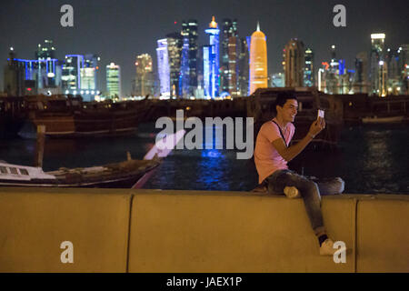 A young man sits on the sea wall of Corniche Promenade making a video call using his mobile phone. In the background is Dhow Harbour and beyond the hi Stock Photo