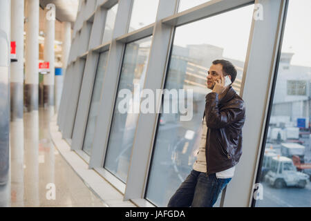 Portrait of young handsome person wearing casual style clothes standing near window in modern airport terminal. Traveler making call using smartphone. Stock Photo