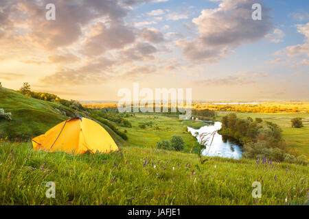 view of tourist tent on green meadow at sunrise or sunset. Camping background with copy space. Tent on the hill near the river. Adventure travel activ Stock Photo