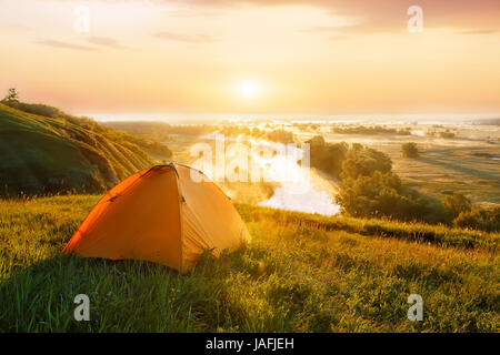 view of tourist tent on green meadow at sunrise or sunset. Camping background. Orange tent on the hill near the river. Adventure travel active lifesty Stock Photo