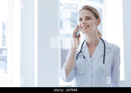 Enjoying conversation. Cheerful delighted young doctor enjoying working hours in the hospital while expressing joy and having conversation on the phon Stock Photo