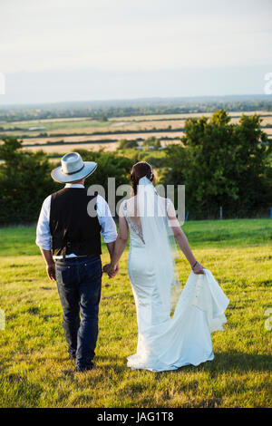 Rear view of bride and groom walking hand in hand across a grassy slope, with a view over the landscape. Stock Photo