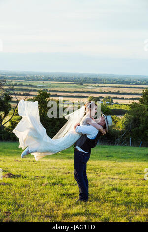 Newlyweds standing in a grass meadow overlooking a rural view, groom hugging and swinging the bride in the air. Stock Photo
