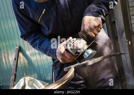 Man standing outdoors working on a metal pitchfork with a hand held electric angle grinder. Stock Photo