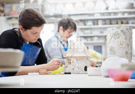 Two women seated at a workbench in a pottery studio, working on decorated clay objects.