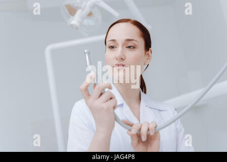 My job. Serious young dentist holding instrument in both hands and looking attentively at it while being ready for work Stock Photo