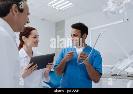 I am only student. Positive girl holding tablet in both hands and standing between two male dentists while looking forward Stock Photo