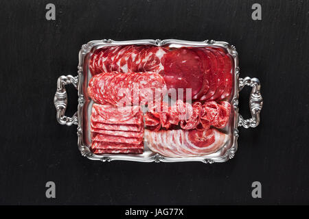 Assortment of different sliced meat snacks. Serving cold Italian appetizers  On a silver tray On black natural stone from slate Stock Photo