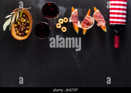 Cold meat cut from different varieties of salami on a dark background with a bottle of red wine and snacks, melons and breadsticks. Italian lunch Stock Photo