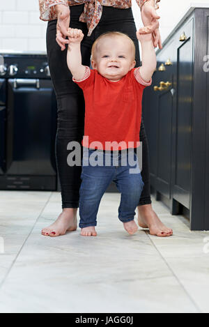 Mother Helping Baby Son To Walk In Kitchen Stock Photo