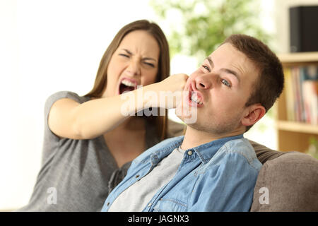 Woman hitting her boyfriend sitting on a couch in the living room in a house interior Stock Photo