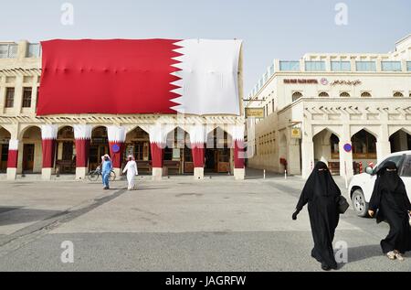Two Qatari women in black abaya robes walking near the Souq under a giant flag of Qatar in the center of Doha Stock Photo