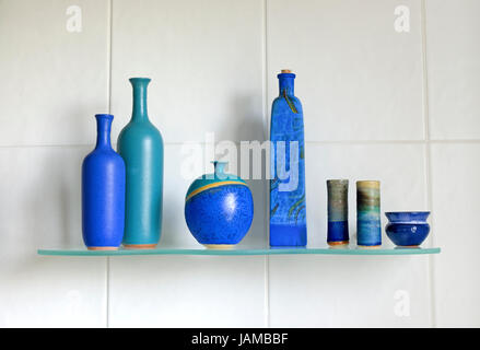 Still life arrangement of beautiful hand made artisan blue artisan pottery on a glass shelf, with a white tiled background. Stock Photo