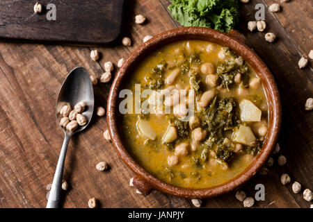 closeup of an earthenware bowl with kale stew with potatoes and chickpeas, on a rustic wooden table sprinkled with some dry chickpeas Stock Photo