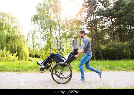 Hipster son running with disabled father in wheelchair at park. Stock Photo