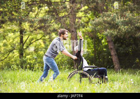 Hipster son walking with disabled father in wheelchair at park. Stock Photo