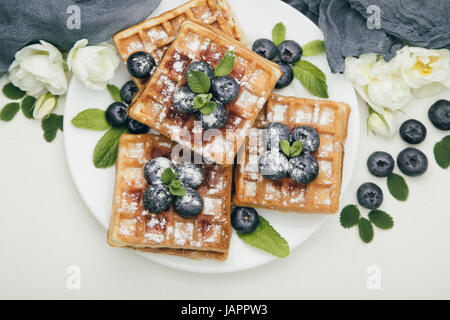 Belgian waffles with fresh blueberries and mint with white flowers on white table. Delicious dessert. Top view Stock Photo