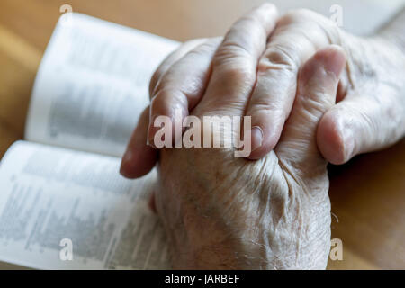 weathered old man's hands clasped in prayer over open Bible Stock Photo