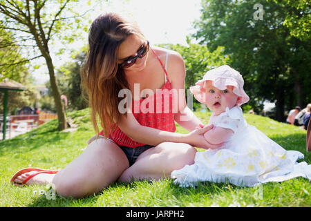 Young mother in her 20s with her six month old baby girl wearing sun hat and dress in a park Stock Photo