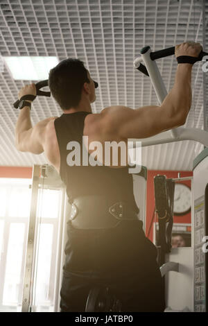Muscular man in a black t-shirt and pants with a weightlifting belt, pulled on the simulator in the sun, back view from below. Vertical photo Stock Photo