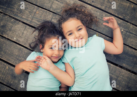 Horizontal shot of two smiling small girls holding hair lying on wooden floor. Stock Photo