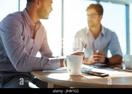 business executives discussing work at office. Closeup of coffee cup with blurred image of two businessmen sitting on table. Stock Photo