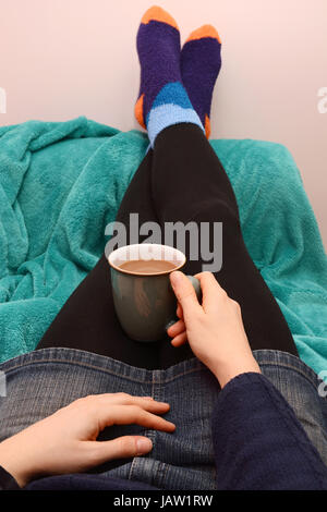 Woman dressed warmly, holding hot coffee or tea, with her feet up on the sofa Stock Photo