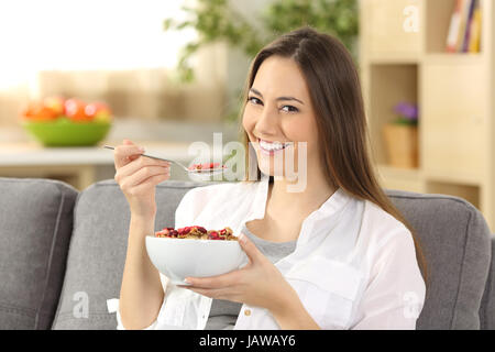 Happy girl eating cereals and looking at you sitting on a sofa in the living room at home Stock Photo