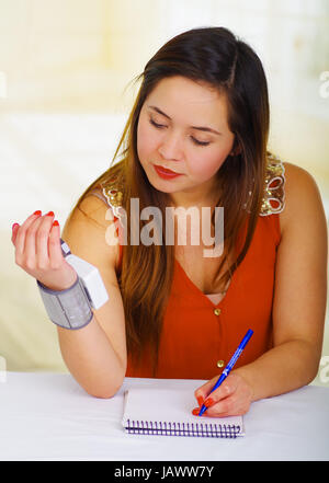 Beautiful young patient using an automatic tensiometer in her arm and taking notes of the results in his notebook, in a doctor consulting room background Stock Photo