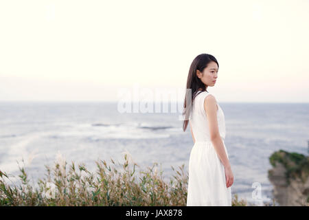 Young Japanese woman in a white dress at a cliff over the sea at sunrise, Chiba, Japan Stock Photo