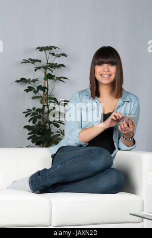 Junge, hübsche Frau sitzt auf dem Sofa und tippt auf ein Smartphone Stock Photo