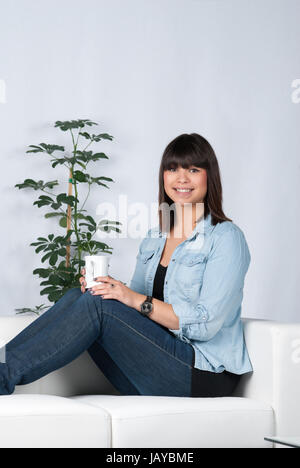Junge, hübsche Frau sitzt auf dem Sofa und hält eine Tasse Kaffee Stock Photo