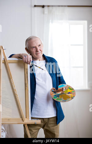 portrait of pensive senior man standing near easel in art workshop Stock Photo