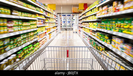 An empty shopping cart between rows of shelves in the supermarket. Stock Photo