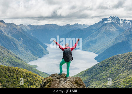 Female hiker is looking at the South Fiord of Lake Te Anau, stretching arms in the air, at back the Southern Alps